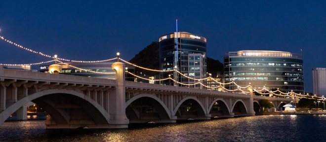 This photo shows a view of Mill Avenue Bridge over the Salt River in Tempe, Arizona, with lights spanning the bridge and buildings in the background.